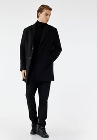 Zwarte wollen jas met ingesneden revers, twee knopen en zijzakken, gecombineerd met een zwarte coltrui en slim-fit broek. Bevat zwarte sneakers.