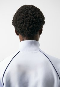 Suéter blanco con un cuello alto, que presenta una tela texturizada y ribetes azul oscuro a lo largo de las costuras, mostrando el cabello rizado del portador desde atrás.