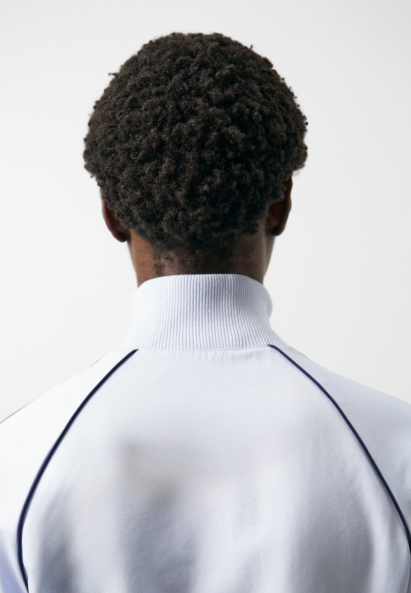 Weißer Pullover mit einem hohen Kragen, aus strukturiertem Stoff und dunkelblauen Paspeln an den Nähten, die die lockigen Haare des Trägers von hinten zeigen.
