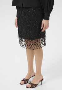 Jupe noire avec des perles florales et une superposition transparente ; associée à des mules noires et marron à motif avec un accent en nœud.