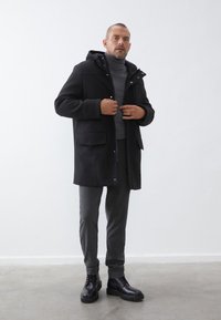 Abrigo negro con capucha y bolsillos frontales, superpuesto a un suéter de cuello alto gris, combinado con pantalones grises y botas negras de cordones sobre un suelo gris.
