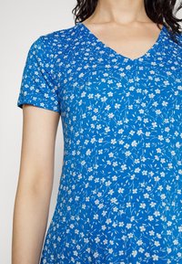 Femme portant une robe bleue à manches courtes en V avec un petit motif floral blanc, montrée de la nuque aux hanches.