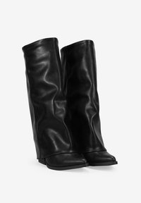 Botas altas de cuero negro con un ligero tacón, punta redondeada y cañas caídas contra un fondo blanco.