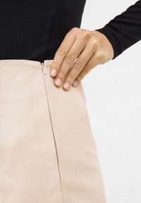 Meruňková kožená sukně s bočním zipem, hladká textura a minimalistický design. Ruka umístěná na oblasti zipu pro úpravu.