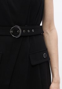 Svart kjole med bredt belte med sirkulær metallspenne, maljer og lappelommer med trykknapper. Glatt stoff med strukturert design.