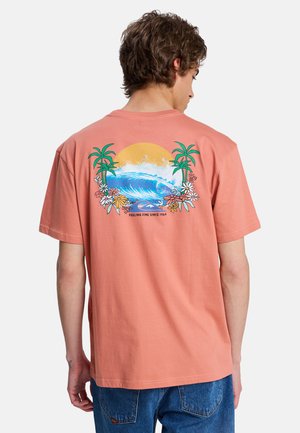 Mężczyzna ubrany w łososiowy t-shirt z falą, słońcem, palmami, kwiatami oraz napisem „Feeling Fine Since 1969” na plecach.
