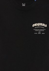 Juodo audinio marškiniai su balta užrašu ant krūtinės „ORIGINALS INTERNATIONAL jungia kūrybą per atstumus“ ir mažu logotipu žemiau.
