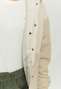 Beige quilted jacket with soft faux-shearling sleeves, snap buttons, and a zip front. White t-shirt underneath, paired with olive green pants.