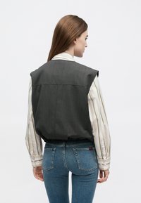 Sivá bez rukávov vesta z textúrovaného materiálu, nosená cez pruhovanú bielu a žltú košeľu, doplnená modrými priliehavými džínsy.