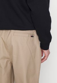 Personne portant un pantalon beige avec une poche arrière et un haut noir à manches longues, main détendue le long du corps sur un fond blanc.