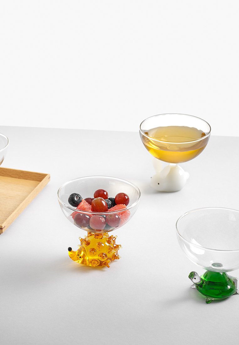 Szklane miseczki na desery z podstawami w kształcie zwierząt; jedna ma przezroczystą krawędź, żółtą podstawę jeża, wypełnioną mieszanką owoców. Pozostałe podstawy to żółw i królik.