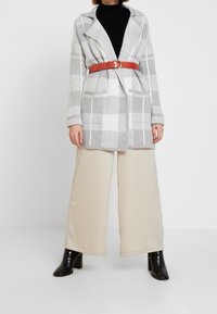 Eine Frau trägt eine graue karierte Strickjacke mit einem roten Gürtel, einen schwarzen Rollkragenpullover, cremefarbene weite Hosen und schwarze Stiefeletten mit Absatz.