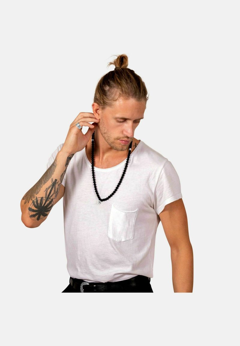 Maglietta bianca a maniche corte con taschino, indossata con una collana di perline nera. Presenta un tatuaggio sull'avambraccio. Lo sfondo è grigio uniforme.