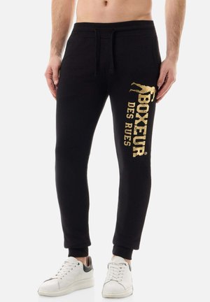 Mann trägt schwarze Jogginghose mit goldenem "Boxeur des Rues"-Text und Silhouette am linken Bein, kombiniert mit weißen Sneakern.