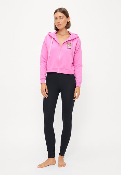 Roza zip-up kratka kapuca z grafiko medveda in logotipom na prsih, v kombinaciji s črnimi leginami. Mehka tkanina, gladka tekstura, prilegajoča zasnova.