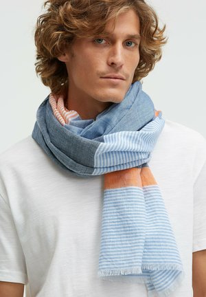 Man with curly hair wearing white t-shirt and a loosely wrapped scarf with blue, orange, and white striped patterns around his neck.