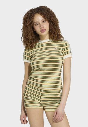Ung kvinde med krøllet hår iført matchende gul, grøn og brun stribet kortærmet skjorte og shorts, stående mod en hvid baggrund.