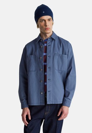 Młody mężczyzna w granatowej czapce, niebieskiej kurtce zapinanej na guziki na bordowej i niebieskiej pasiastym koszuli oraz ciemnoniebieskich dżinsach, stojący na białym tle.