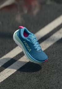 Baskets de course bleues avec une tige en maille texturée, semelle superposée verte et blanche, languette au talon rose et logo HOKA bien en évidence.