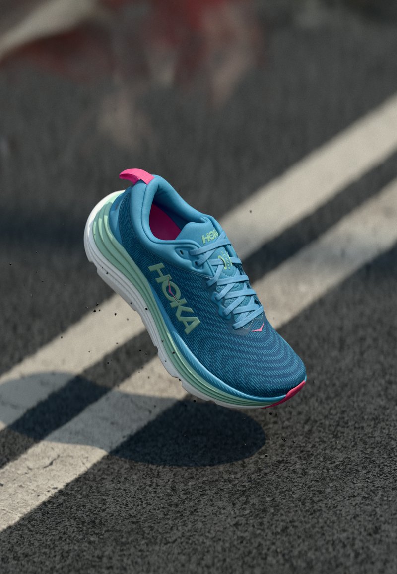 Baskets de course bleues avec une tige en maille texturée, semelle superposée verte et blanche, languette au talon rose et logo HOKA bien en évidence.