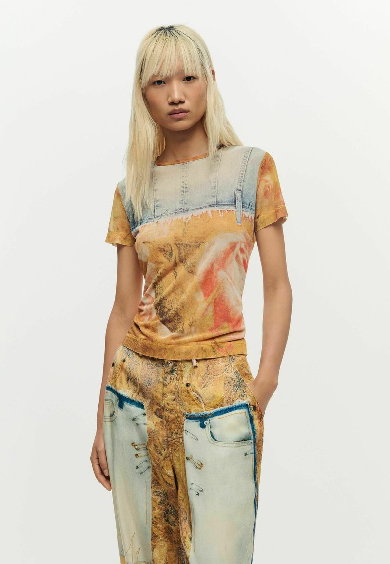 Kobieta o prostych blond włosach, ubrana w dopasowany krótki rękaw i spodnie z pomarańczowym i dżinsowym wzorem, z rękami w kieszeniach.