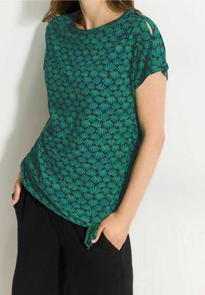 Green short-sleeved top with a floral pattern, featuring cut-out shoulder details and a loose fit, paired with black pants.