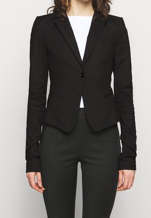 Femme portant un blazer noir ajusté avec un seul bouton, associé à un pantalon noir et un haut blanc en dessous.