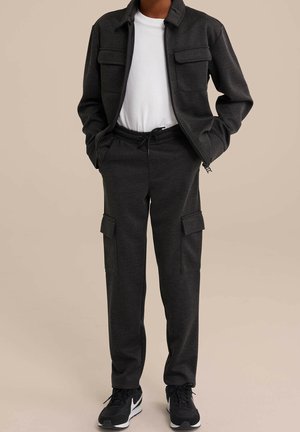 Osoba v černých cargo kalhotách a bundě přes bílé tričko, stojící s rukama v kapsách, viditelné černobílé tenisky.