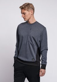 Mörkgrå collegetröja med en texturerad yta, med en ficka framtill som går att dra upp med dragkedja och svarta detaljer på sidorna och ärmarna.