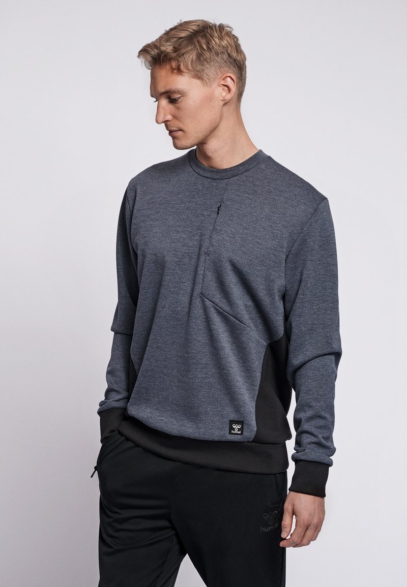 Mörkgrå collegetröja med en texturerad yta, med en ficka framtill som går att dra upp med dragkedja och svarta detaljer på sidorna och ärmarna.