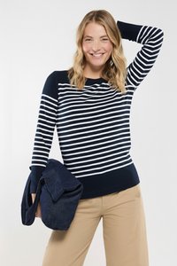 Gestreifter Pullover in Marineblau und Weiß mit langen Ärmeln, einem runden Halsausschnitt und einem einfarbigen Marineblau-Saum. Eine Denim-Tasche wird in einer Hand gehalten. Hosen in neutralen Farben.