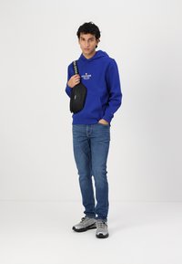 Sweatshirt à capuche bleu avec poche avant et logo blanc, associé à un jean bleu et des sneakers grises. Un petit sac crossbody noir est porté.