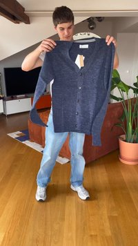 Osoba držící modrý kardigan s knoflíky a štítky v obývacím pokoji s dřevěnou podlahou a květináčem.