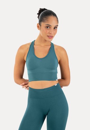 Kobieta w turkusowym bezszwowym sportowym crop topie i legginsach, stojąca z rękami za plecami, patrząca w obiektyw na jednolitym tle.