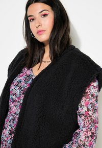 Schwarze Fleece-Weste mit einer strukturierten, flauschigen Oberfläche, die über einem blumigen Druckkleid in Pink-, Blau- und Weißtönen getragen wird.