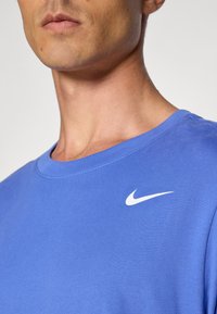 Primer plano de una persona vistiendo una camiseta azul de Nike con el logo en forma de swoosh blanco en el lado izquierdo del pecho, contra un fondo liso.