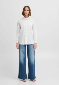 Chemise blanche à manches longues avec une poche poitrine, associée à un jean bleu clair à jambes larges avec un motif décoloré et des ourlets effilochés.