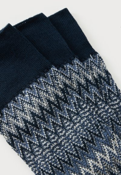 Trois chaussettes tricotées avec des bord-côtes bleu foncé et un motif en zigzag dans des tons de bleu, noir et blanc sur la zone du pied.