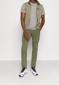 Olivgröna cargobyxor i kombination med en grå t-shirt och en beige ärmlös jacka. Vita sneakers med svarta detaljer fulländar looken.