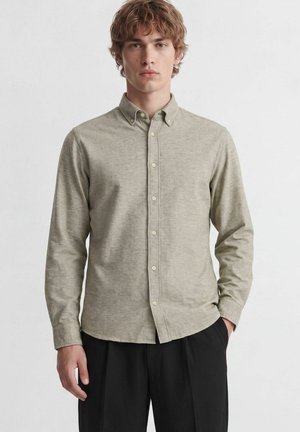 Junger Mann mit lockigem Haar, der ein hellgraues Hemd mit Knöpfen und schwarze Hose trägt, steht mit einer Hand in der Tasche vor einem einfarbigen Hintergrund.