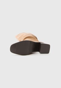 Semelle de chaussure plate face avant avec une tige de botte beige floue et un petit talon carré sur un fond blanc uni.
