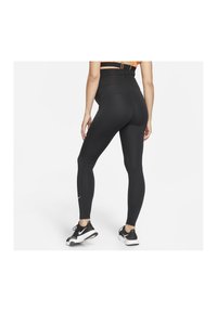 Donna che indossa leggings neri a vita alta e sneakers Nike bianco e nero, in piedi con una gamba leggermente piegata su uno sfondo bianco semplice.