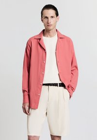 Hellrosa Hemd mit Knopfleiste und lockerem Schnitt, kombiniert mit einem cremefarbenen T-Shirt und beigen Shorts. Verfügt über einen geflochtenen schwarzen Gürtel. Glatte Textur.