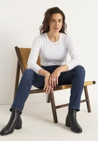 Vit långärmad topp, mörkblå jeans, svarta ankelskor, sittande på en trästol med vävd sits. Neutral bakgrund, med fokus på kläder.