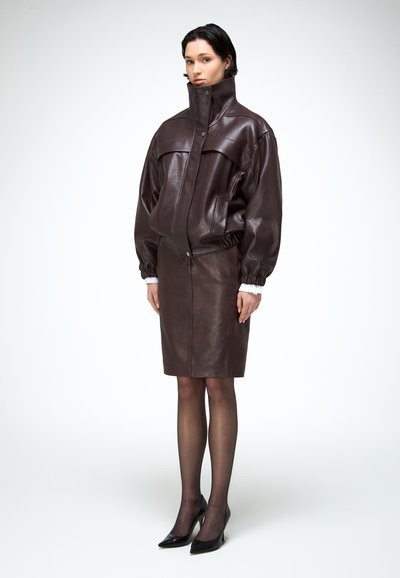 Nő sötétbarna bőrkabátot visel magas gallérral, hozzá illő bőr szoknyát, áttetsző fekete harisnyát és fekete hegyes sarkú cipőt, fehér háttér előtt állva.