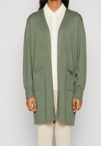 Person som bär en öppen sagegrön kofta med två framfickor över en vit skjorta med knappar och krämfärgade ribbade byxor.