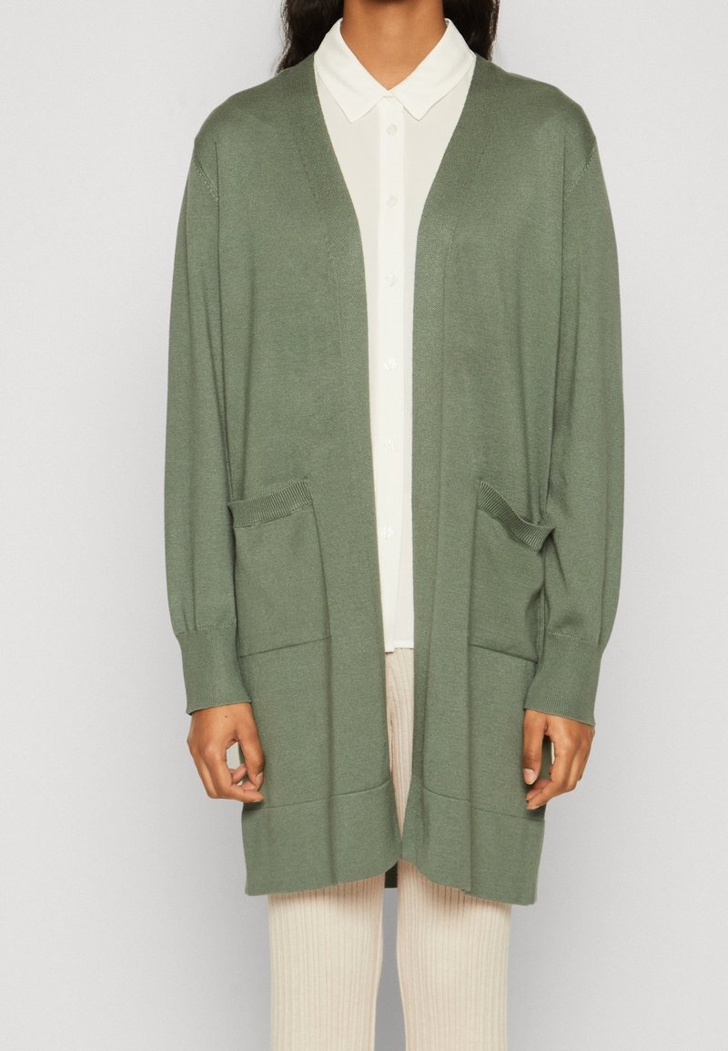 Person som bär en öppen sagegrön kofta med två framfickor över en vit skjorta med knappar och krämfärgade ribbade byxor.