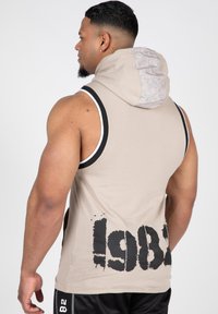 Beige mouwloze hoodie met een contrasterende witte en zwarte rand. Heeft een gemêleerde grijze capuchon en een grote zwarte "1982" print op de achterkant.