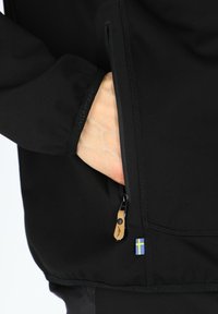 Black jacket featuring a zipped pocket, textured fabric, and a Swedish flag detail near the zipper. Close-up of a hand in the pocket.