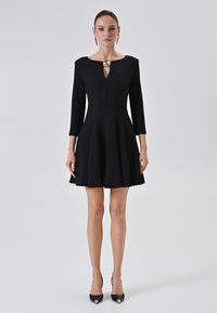 Schwarzes, tailliertes Kleid mit drei Viertelärmeln, ausgestelltem Rock und einem Schlüssellochausschnitt. Goldfarbene florale Akzentuierung am Kragen. Trägt schwarze High Heels.